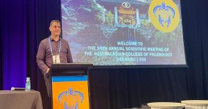 A man stands at a podium on stage next to a table with a bottle of water. Behind him, a screen displays "24th Annual ACP Scientific Meeting of the Australasian College of Phlebology, Denarau, Fiji,"