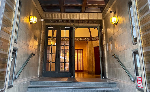 A building entrance with glass double doors, flanked by wall-mounted lanterns, and handrails on both sides of the steps leading to the door.