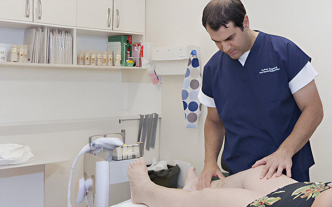 A healthcare professional examines a patient's leg in a clinical setting. Medical supplies are visible on shelves in the background.