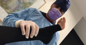 A medical professional in scrubs carefully examines a patient's leg, covered in a black compression stocking.