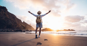 Person with backpack and hat stands on a beach, with arms raised, facing the ocean and sunset. Rocky cliffs are in the background.