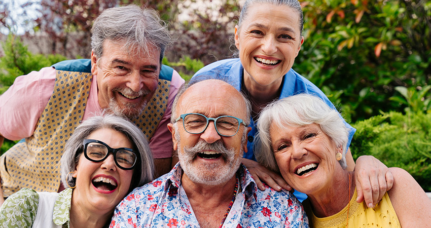 Five older adults smiling outdoors in a garden, three standing behind and two sitting in front. They appear happy and relaxed.